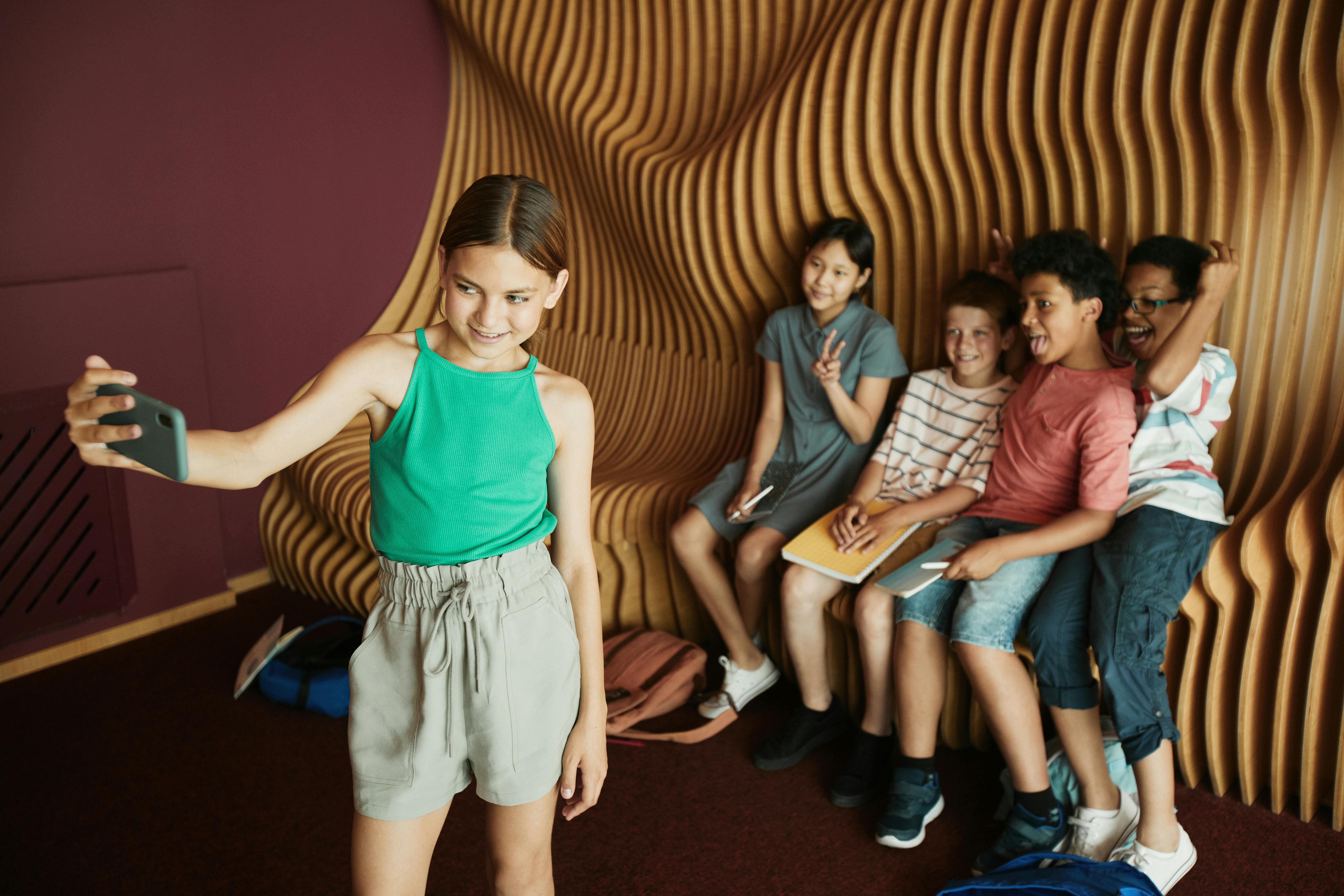 Young girl wearing a green tank top and khaki shorts taking a selfie, standing in front of a group of 3 young boys who are posing on a decorative bench